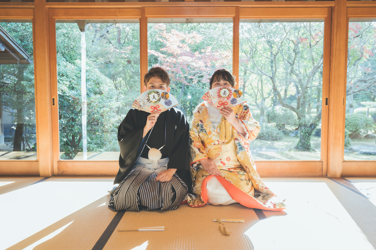 和室で新郎と新婦が扇子を口元に当てて口を隠している写真
