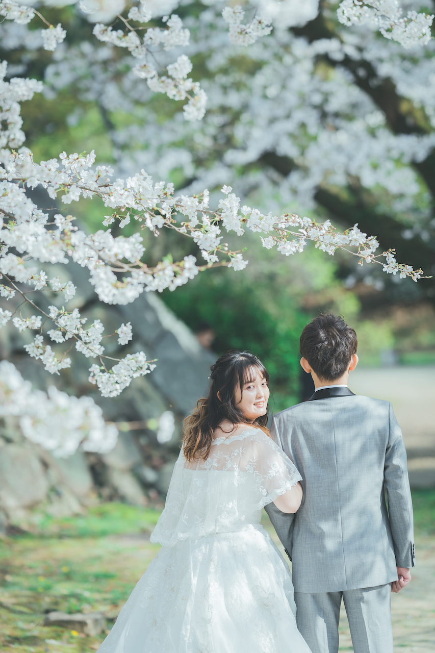 新郎と新婦が桜の下に立っており、新婦がこちらを振り向いている写真