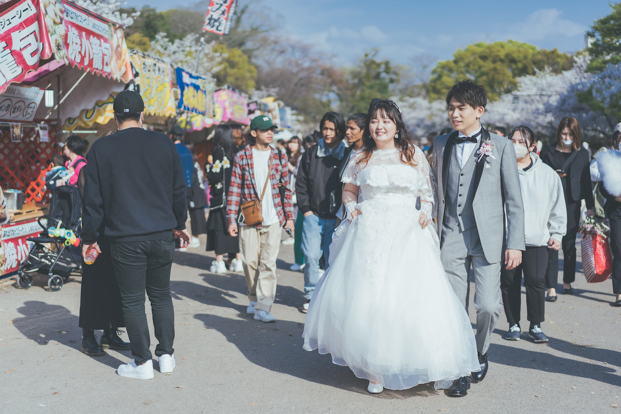 新郎と新婦がタキシードとウェディングドレスで桜まつりの屋台通りを練り歩いている写真